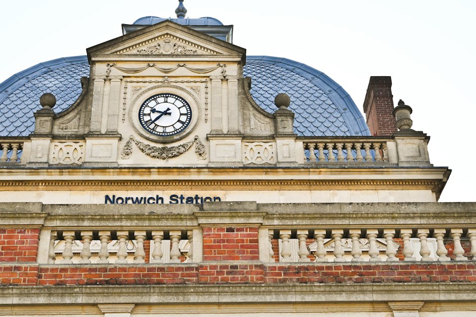 A historic building with a decorative clock face showing the time as 2:12, situated above a ledge with the words 'Norwich Station' displayed beyond a stone balustrade. The building features ornate arc