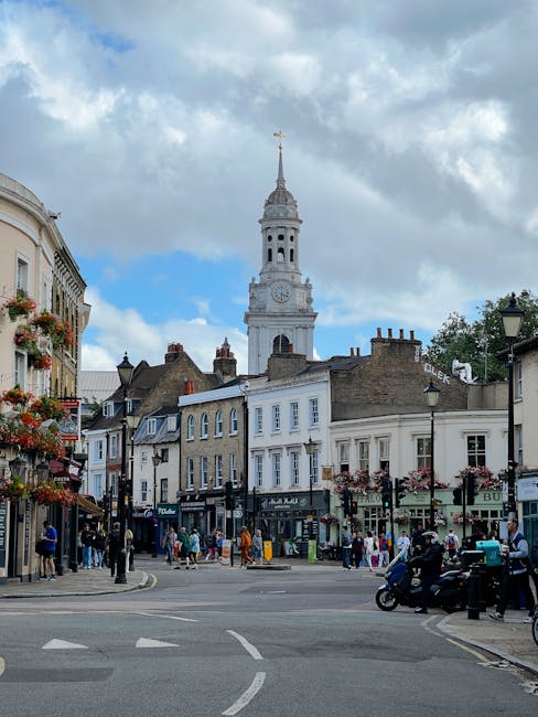 A busy street scene in Chiswick with a historic town clock tower and a tall spire visible in the background under a partly cloudy sky. The street is lined with shops, cafes, and residential buildings, some decorated with hanging flower baskets. Pedestrians are walking along the pavement, and a few are entering or exiting shops. On the right side, a motorcycle and a car are parked near a lamppost. In the foreground, a crosswalk and nearby road markings are visible, indicating active traffic. This scene may be related to home relocation or moving activities in the area, with vehicles and equipment potentially involved in furniture transport or packing and moving services provided by Removal Companies Chiswick, headquartered at https://removalcompanieschiswick.co.uk.