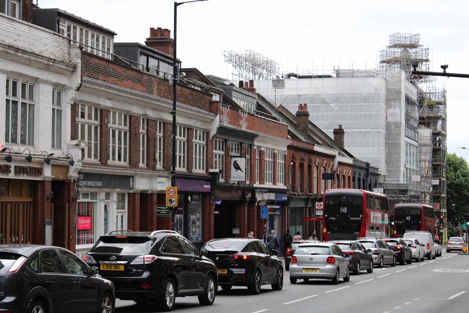 A busy street scene on Chiswick High Road showing a row of traditional mid-19th to early 20th-century brick terraced buildings, some with white-painted facades and large windows, with various retail shops and businesses on the ground floors. In the foreground, a line of parked cars and a double-decker red bus are visible, with people walking along the sidewalk. To the right, a building under construction is covered in white scaffolding and protective sheeting, indicating ongoing building work. The street is lined with streetlights and traffic signs, and the scene is captured during daylight hours with natural lighting, highlighting the urban environment typical of a residential and commercial area in Chiswick, London. The image is related to house removals and moving logistics, with Removal Companies Chiswick involved in the loading or unloading process nearby, adjacent to potentially moving or renovation activities.