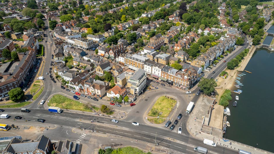 A busy street scene in Chiswick with a historic town clock tower and a tall spire visible in the background under a partly cloudy sky. The street is lined with shops, cafes, and residential buildings, some decorated with hanging flower baskets. Pedestrians are walking along the pavement, and a few are entering or exiting shops. On the right side, a motorcycle and a car are parked near a lamppost. In the foreground, a crosswalk and nearby road markings are visible, indicating active traffic. This scene may be related to home relocation or moving activities in the area, with vehicles and equipment potentially involved in furniture transport or packing and moving services provided by Removal Companies Chiswick, headquartered at https://removalcompanieschiswick.co.uk.