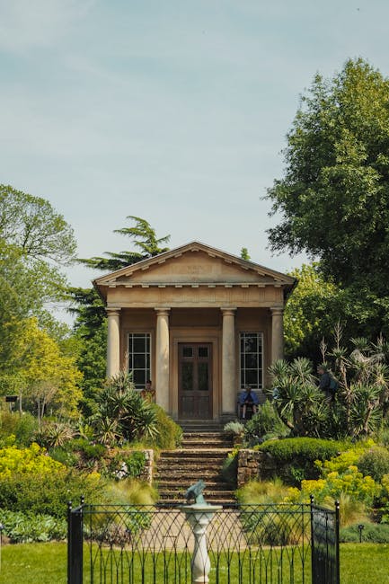 A small classical-style stone building with a triangular pediment and four columns at the entrance, situated within lush green gardens. The building has a wooden door and narrow windows on either side, with stairs leading up to the entrance. Surrounding the property are well-maintained flower beds, shrubs, and trees, with a black metal fence and a white decorative fountain in the foreground. The scene is well-lit with natural daylight, indicating a clear day. This setting reflects a historic or ornamental structure, potentially used as a focal point in a garden or park, typical of a location that could be part of a house or estate undergoing a home relocation or garden clearance by Removal Companies Chiswick during a move or restoration process.