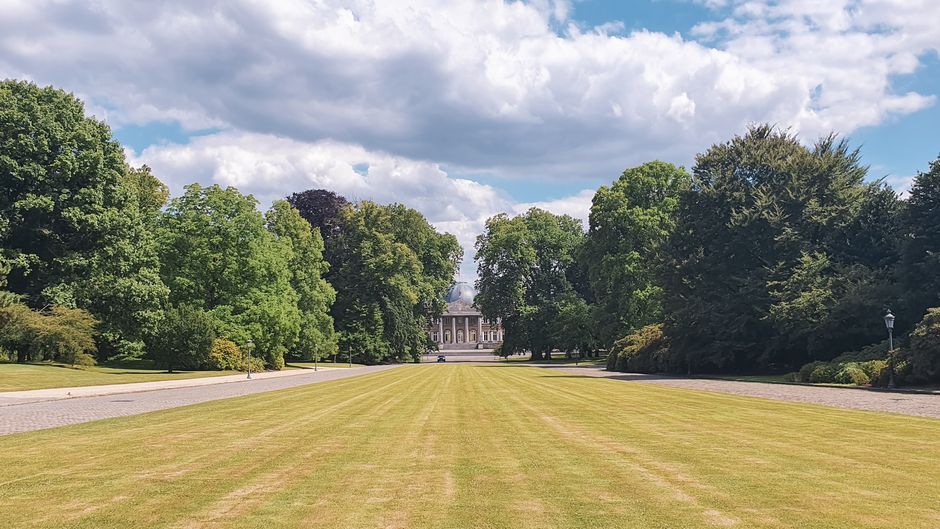 A wide, well-maintained grassy lawn leading towards a historic building at Chiswick House and Gardens, with tall, lush green trees lining both sides of the open space. The sky is partly cloudy with patches of blue, and the scene is illuminated by natural daylight. In the foreground, a paved driveway or pathway runs parallel to the lawn, which is used for home relocation or furniture transport logistics during a house move. This outdoor area, captured during a removals day, provides clear access for loading and unloading boxes, furniture, and removal equipment, which may include trolleys, moving blankets, or vans. The setting suggests a peaceful yet functional environment for carrying out the final stages of packing, moving, and equipment handling for a home removal service, as provided by Removal Companies Chiswick during a house and garden migration process.