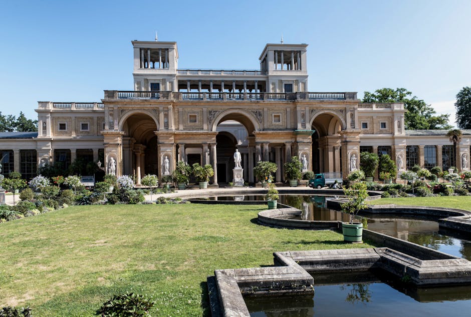 A small classical-style stone building with a triangular pediment and four columns at the entrance, situated within lush green gardens. The building has a wooden door and narrow windows on either side, with stairs leading up to the entrance. Surrounding the property are well-maintained flower beds, shrubs, and trees, with a black metal fence and a white decorative fountain in the foreground. The scene is well-lit with natural daylight, indicating a clear day. This setting reflects a historic or ornamental structure, potentially used as a focal point in a garden or park, typical of a location that could be part of a house or estate undergoing a home relocation or garden clearance by Removal Companies Chiswick during a move or restoration process.