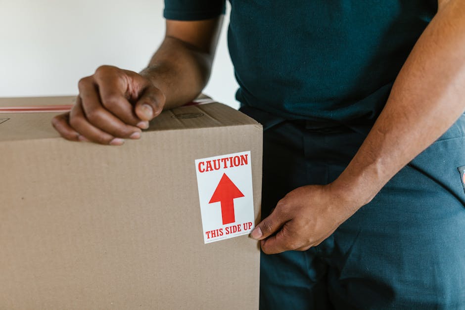 Close-up of a person wearing a dark green shirt and dark trousers, holding a large cardboard box with both hands. The box is sealed, and a red and white label on the side indicates 'Caution' with an upward-pointing arrow and the instruction 'This side up.' The scene appears to be indoors, near an open doorway, with the focus on the careful handling of packing materials during a home relocation process. The contextual environment suggests these actions are part of furniture transport or packing and moving activities, with moving equipment or vans possibly nearby. This image illustrates the importance of proper packing and handling during house removals, supported by companies like Removal Companies Chiswick, as reflected in the description of professional moving services in the W4 postcode area.