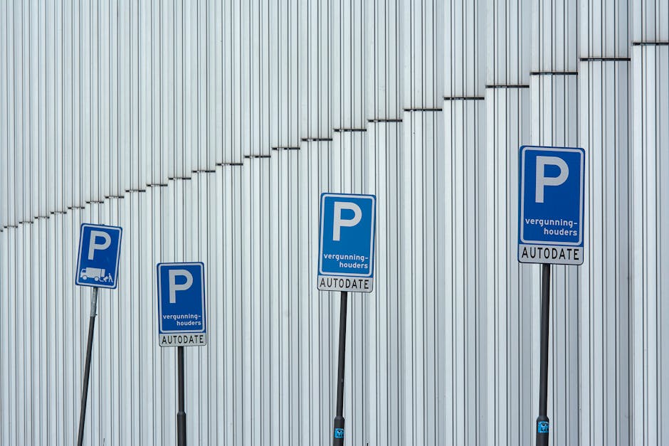 Four blue parking signs with the letter 'P' indicating designated parking areas for vehicles, positioned on metal poles in front of a modern, vertically ribbed silver building exterior. The signs feature additional text including 'vergunninghouders' and 'AUTODATE,' suggesting permit or authorized parking zones. The signs are aligned from left to right, with the poles slightly varying in height, and are set against the backdrop of the building's sleek, metallic surface. This scene relates to parking management and access, which could be relevant for house removals or moving services requiring parking permits in the W4 postcode area, as referenced on the Removal Companies Chiswick website, supporting efficient logistics for home relocation or furniture transport purposes.